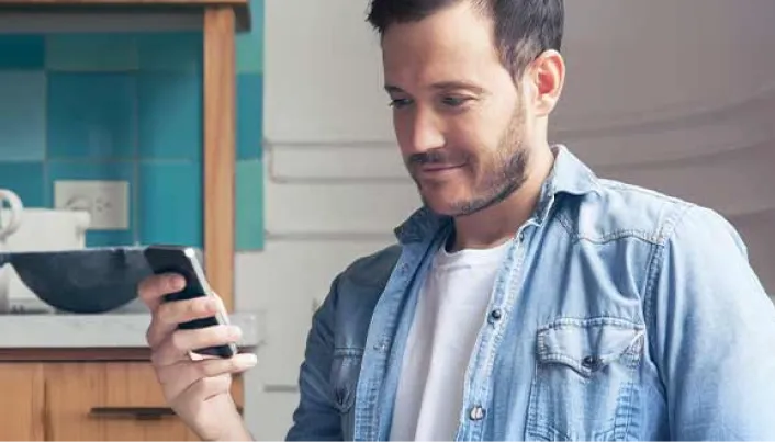 Persona con camisa de mezclilla sosteniendo un teléfono móvil y mirando la pantalla mientras se encuentra en un espacio interior de hogar, con una cocina visible al fondo.