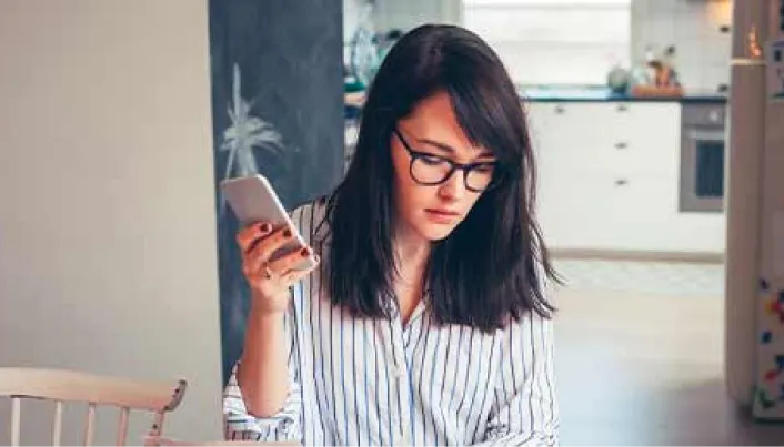 Persona utilizando un teléfono móvil desde el hogar, con una cocina al fondo, en un entorno cotidiano de consulta y gestión digital.