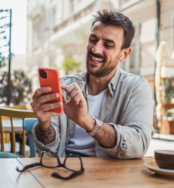 Un hombre sonriente revisando su celular en una cafetería con sus lentes sobre la mesa.