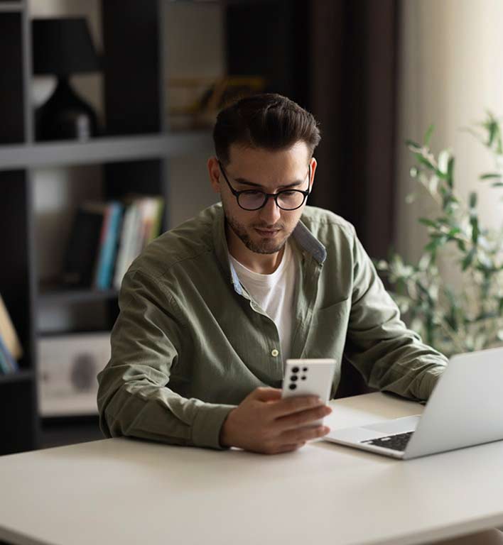 Un hombre sentado frente a un escritorio revisando su celular junto a una laptop abierta.