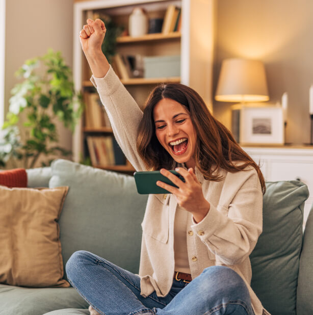Una mujer sentada en un sillón celebrando mientras revisa en su celular el crecimiento de sus ahorros.