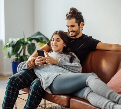 Una pareja en un sillón, ella acostada y recargada sobre él, revisa juntos su celular.